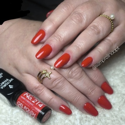 Mãos femininas com unhas pintadas de vermelho vivo e anéis dourados seguras um frasco de verniz gel vermelho