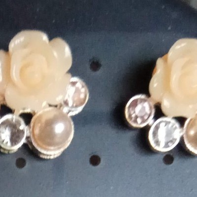 Brincos com flores creme, pérolas e pedras brilhantes em suporte escuro.