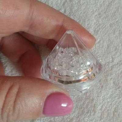 Recipiente transparente em forma de diamante com pequenas esferas, segurado por mão com unha rosa