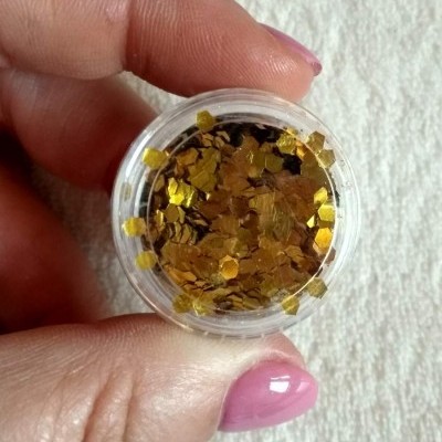 Frasco com glitter dourado segurado por mão com unhas pintadas cor rosa