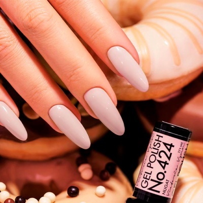 Mãos com unhas compridas cor bege claro e verniz gel, donuts ao fundo, frasco pequeno de verniz gel rosa claro.