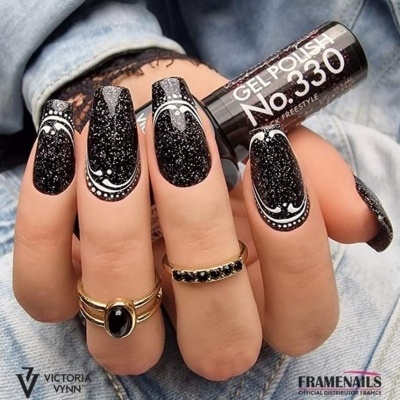 Unhas com verniz gel preto brilhoso e desenhos brancos, mão com anéis dourados segurando verniz gel.