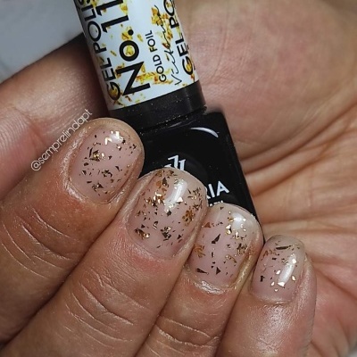 Unhas com verniz gel transparente e flocos dourados segurando frasco de verniz gel