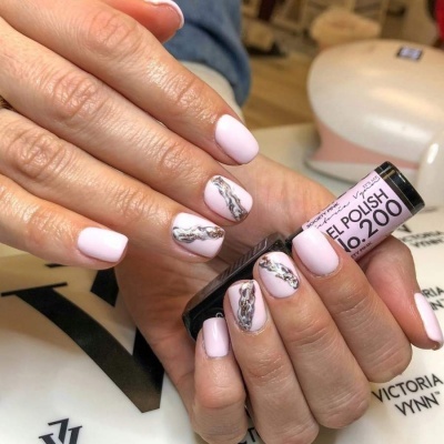 Unhas com gel polish rosa claro e padrão mármore cinza, com frasco de verniz gel na mão e dispositivo UV ao fundo