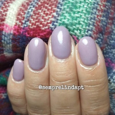 Unhas com verniz lilás opaco sobre fundo de tecido colorido com padrão xadrez.