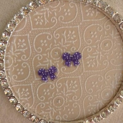 Brincos laço roxo com pontos brancos em moldura circular com pedras brilhantes sobre fundo creme texturizado.