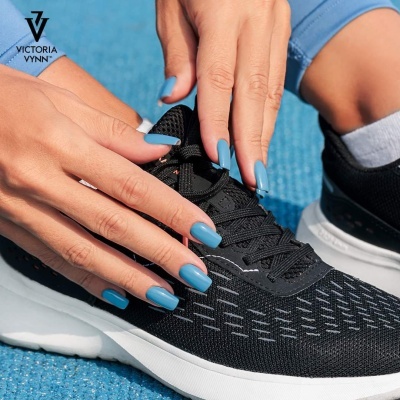 Unhas azuis claras foscas segurando um ténis desportivo preto com sola branca sobre fundo azul texturizado