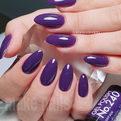 Unhas com verniz gel roxo amêndoa segurando pedra facetada e frasco de verniz gel