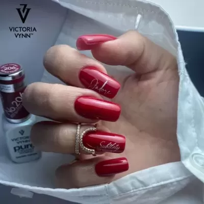 Unhas postiças vermelhas com texto e garrafa de verniz Victoria Vynn ao fundo.