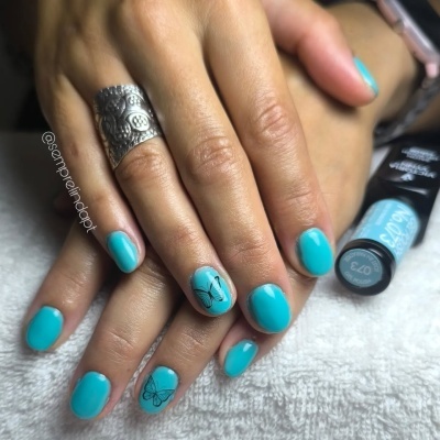 Unhas pintadas de azul turquesa com desenhos pretos de borboleta e nota musical, anel prateado, verniz ao fundo