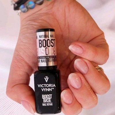 Frasco de verniz para unhas Victoria Vynn Boost Base Nail Repair numa mão feminina com unhas naturais e nude brilhante