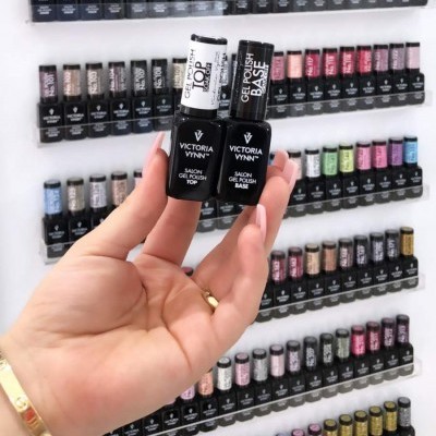 Mão segurando dois frascos de verniz gel Victoria Vynn com expositor de vários vernizes coloridos ao fundo
