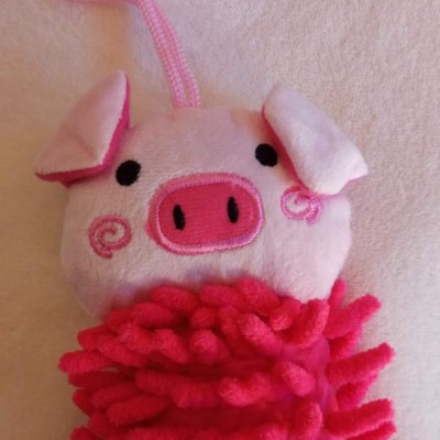 Boneco de pelúcia porquinho rosa com focinho rosa e preto, corpo texturizado em rosa escuro, pendurado sobre fundo claro