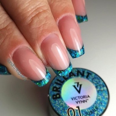 Unhas com ponta azul cintilante e recipiente com rótulo 'BRILLIANTES VICTORIA VYNN 01'