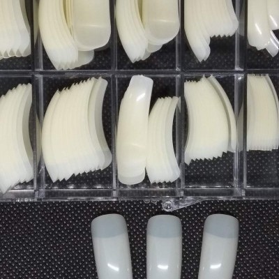 Unhas postiças brancas organizadas em caixa transparente com três unhas expostas sobre fundo preto