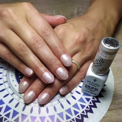 Mãos femininas com unhas pintadas em verniz rosa brilhante e frasco de verniz Victoria Vynn Pure ao lado.