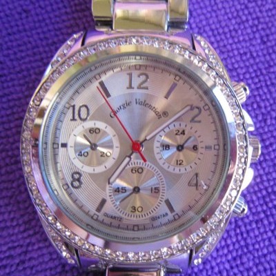 Relógio de pulso prateado com pedras e mostrador branco sobre tecido roxo