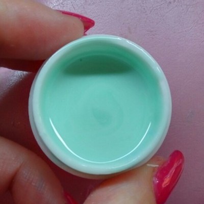 Recipiente branco com líquido verde-água segurado por mãos com unhas vermelhas sobre fundo rosa