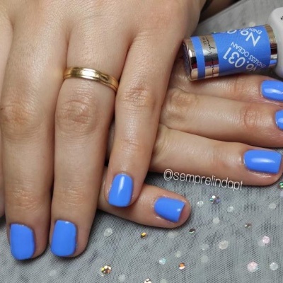 Mãos com unhas pintadas de azul brillante e anel dourado, frascos de verniz ao fundo