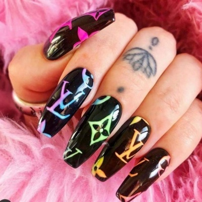 Unhas pretas brilhantes com padrões coloridos e tatuagens na mão sobre fundo rosa peludo