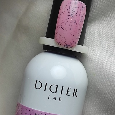 Frasco de verniz gel branco com tampa preta da marca DIDIER LAB e unha artificial rosa com manchas pretas.
