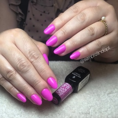 Mãos com unhas cor de rosa vibrante e verniz brilhante, com frasco de verniz ao fundo