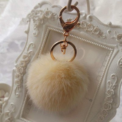 Chaveiro com bola de pelo creme e metais dourados pendurado em suporte branco decorativo