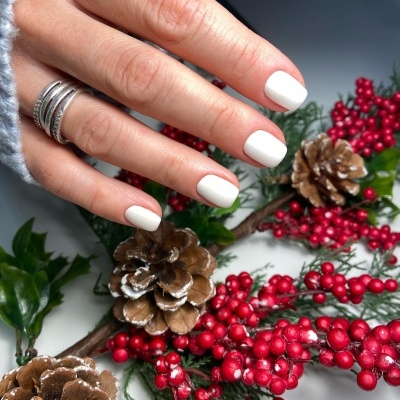 Mão feminina com unhas brancas, anéis prateados e decoração de pinhas e bagas vermelhas