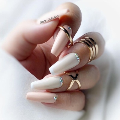 Unhas compridas com esmalte nude e branco e cristais decorativos, com vários anéis dourados nos dedos