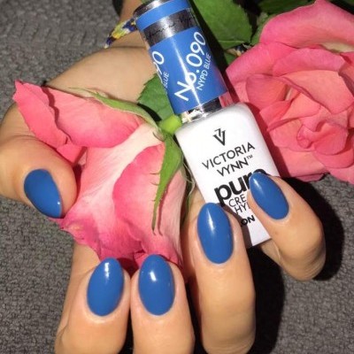 Mãos femininas com unhas pintadas de verniz azul opaco segurando frasco de verniz Victoria Vynn e rosas cor-de-rosa