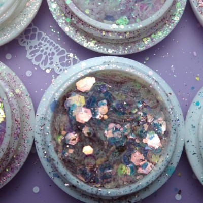 Potes pequenos de plástico branco com glitter holográfico colorido em superfície roxa com padrão de renda.
