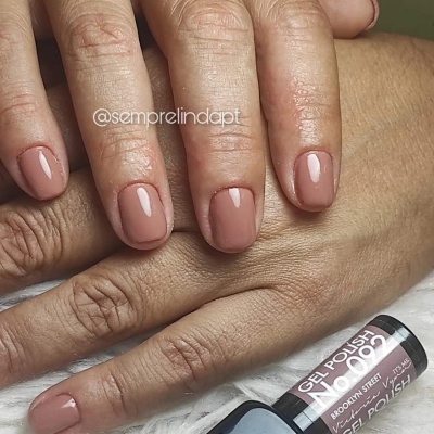 Unhas com verniz castanho claro em gel e frasco de verniz gel no fundo.