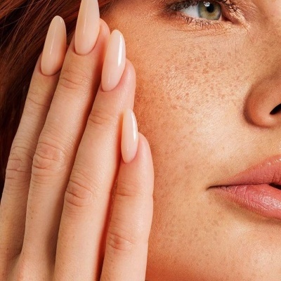 Mão com unhas nude longas tocando rosto de mulher ruiva com sardas