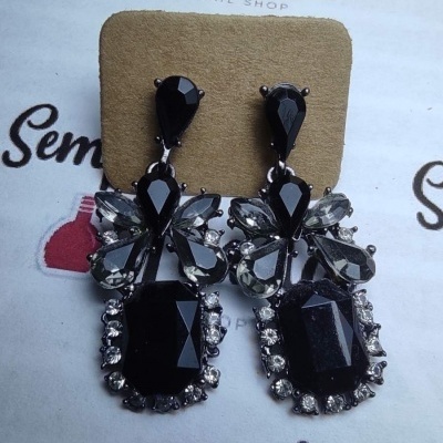 Brincos pretos grandes com pedras facetadas e pedras transparentes, apresentados em suporte castanho com fundo com texto