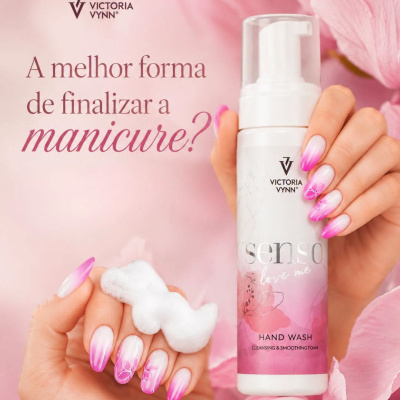Frasco de espuma para mãos com unhas decoradas em rosa e branco e flores cor-de-rosa ao fundo.