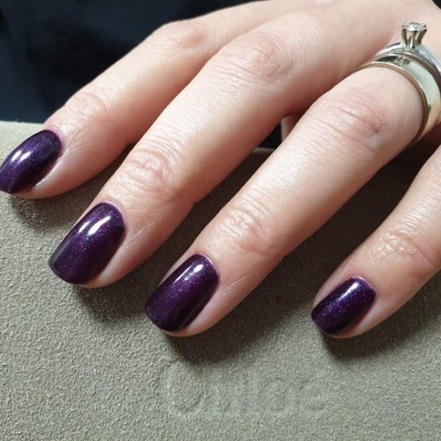 Unhas curtas pintadas de roxo escuro brilhante com dois anéis prateados numa mão sobre superfície cinza.