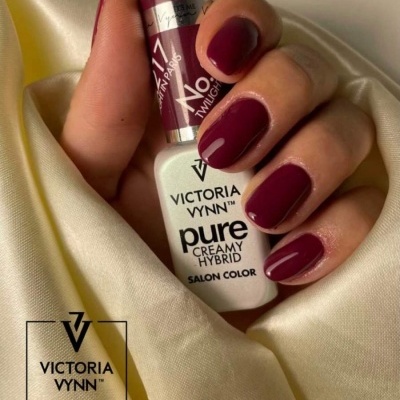 Mão com unhas cor vinho a segurar verniz Victoria Vynn sobre tecido amarelo claro