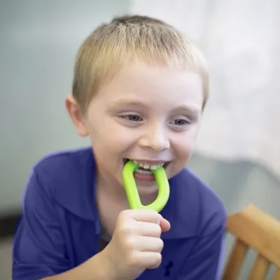 Mordedor ARK's Grabber® liso Menino loiro com peça de plástico verde em forma de D na boca, sentado numa cadeira de madeira