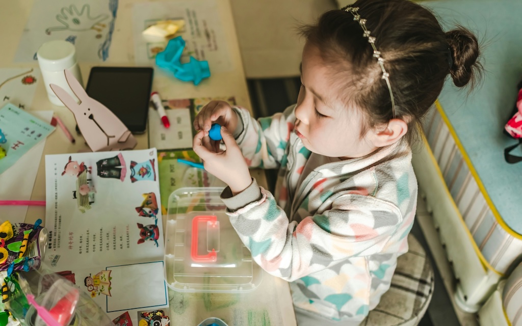 Criança a brincar com massa de modelar azul numa mesa com materiais educativos e acessórios coloridos