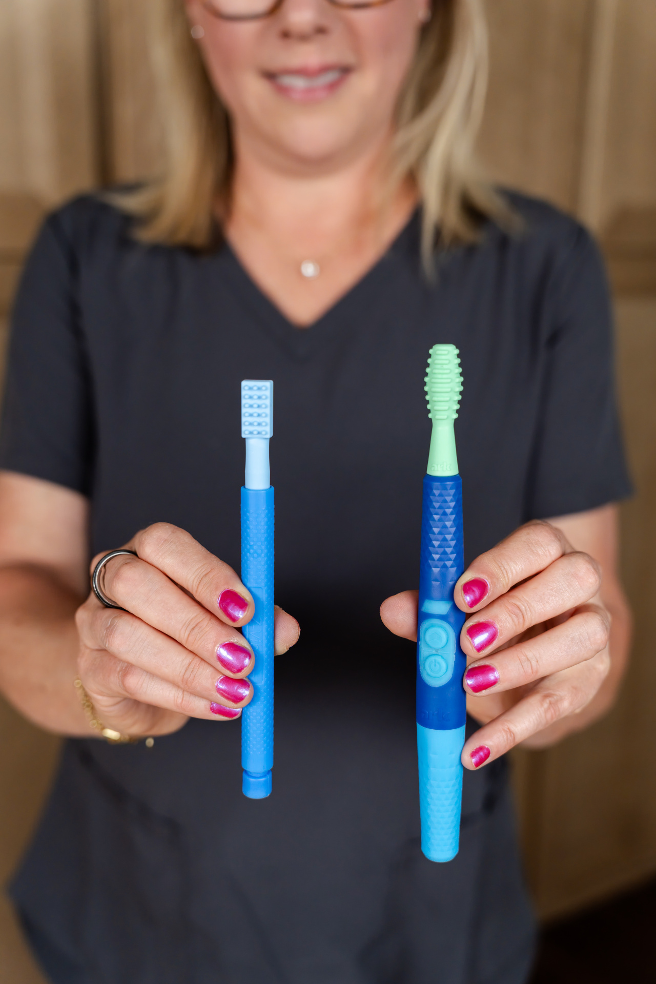 Z-Vibe® Plus da ARK Mulher a segurar duas escovas de dentes de cores azul e verde claro com cerdas de silicone
