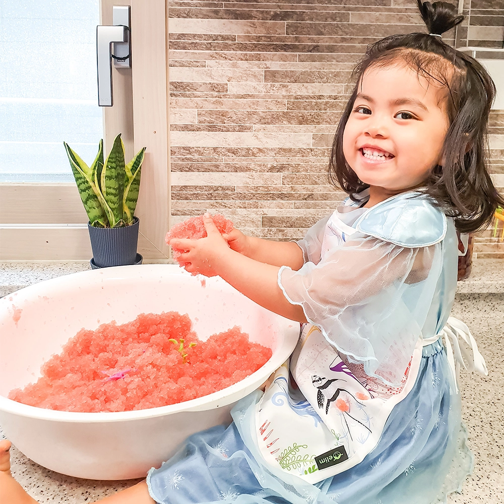 Gelli Play - Gelatina para exploração sensorial Menina sorridente com vestido azul e avental branco junto a bacia branca com pedaços cor-de-rosa e planta num vaso azul