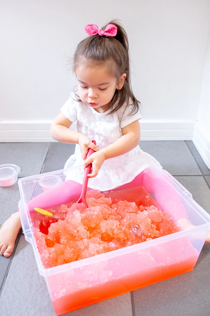 Gelli Play - Gelatina para exploração sensorial Menina com vestido branco mexendo gelo cor-de-rosa numa caixa transparente