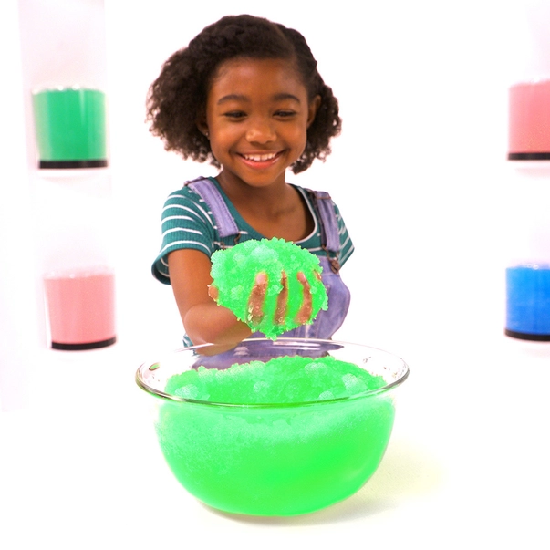Gelli Play - Gelatina para exploração sensorial Criança a brincar com slime verde num recipiente de vidro, com frascos coloridos na parede ao fundo.