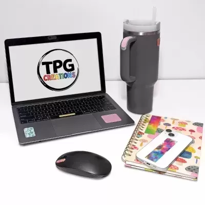 Laptop preto com logotipo TPG CREATIONS, copo térmico cinza com detalhes rosa, rato preto, caderno colorido, smartphone colorido em superfície branca.