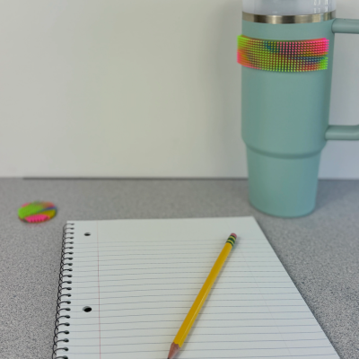 Caderno com lápis numa mesa e caneca térmica azul com faixa de silicone colorida