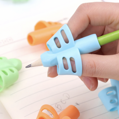 Mão a segurar lápis verde com afinador de escrita azul de silicone sobre papel pautado com outros afinadores coloridos ao fundo