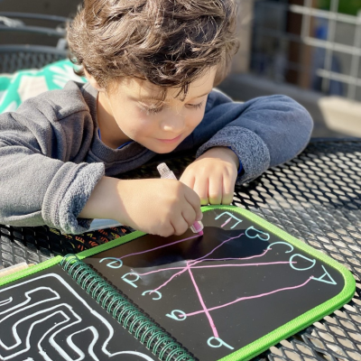 Criança a desenhar num quadro magnético preto com bordas verdes numa mesa metálica preta.