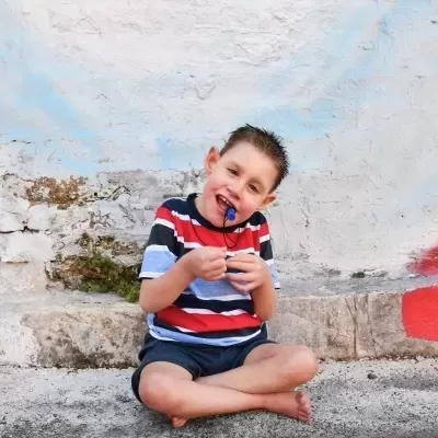 Camisola às riscas com calções, criança sentada no chão em pedra em frente a parede branca