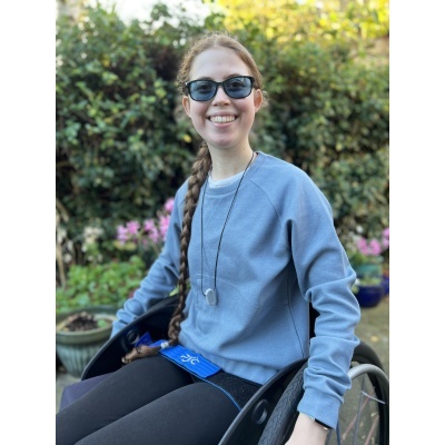 Pessoa usando suéter azul claro e calças pretas, sentada em cadeira de rodas num jardim