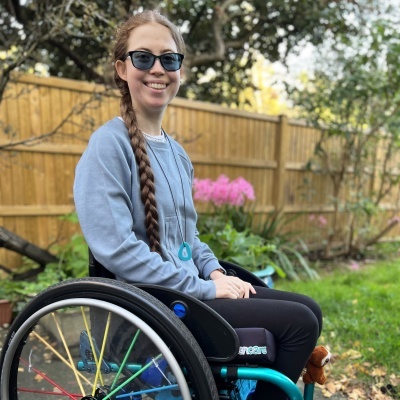 Mulher com óculos numa cadeira de rodas num jardim com cerca de madeira e flores.